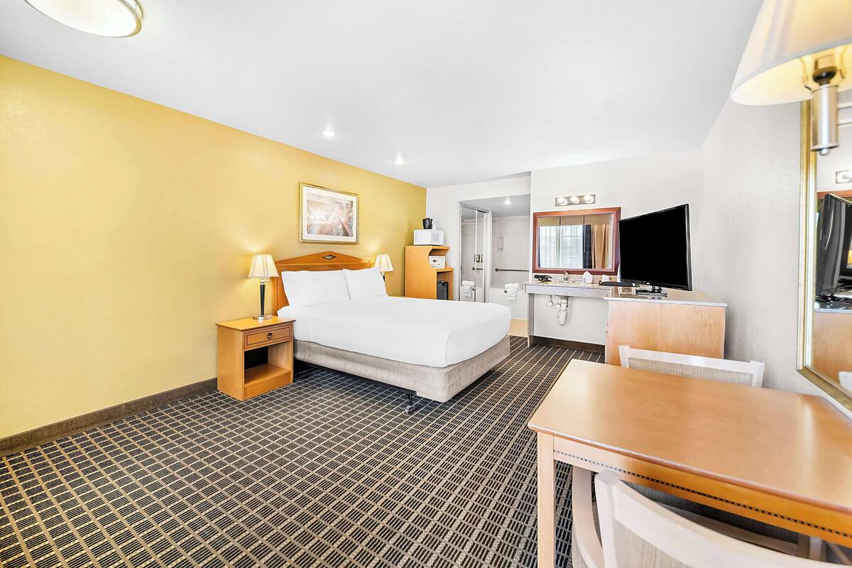 A hotel room with a bed, bedside tables, a desk, a chair, a television, and a bathroom with a mirror and sink.