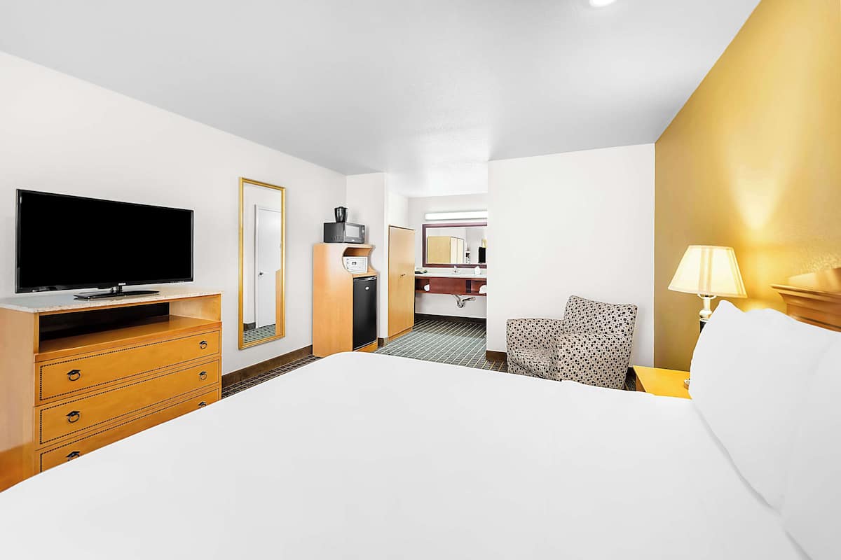 A hotel room with a flat-screen TV, a wooden dresser, a bed, a chair, a bedside table with a lamp, and a bathroom visible through an open door.
