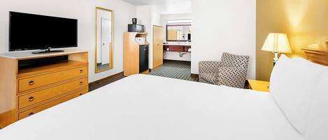 A hotel room with a flat-screen TV, a wooden dresser, a bed, a chair, a bedside table with a lamp, and a bathroom visible through an open door.
