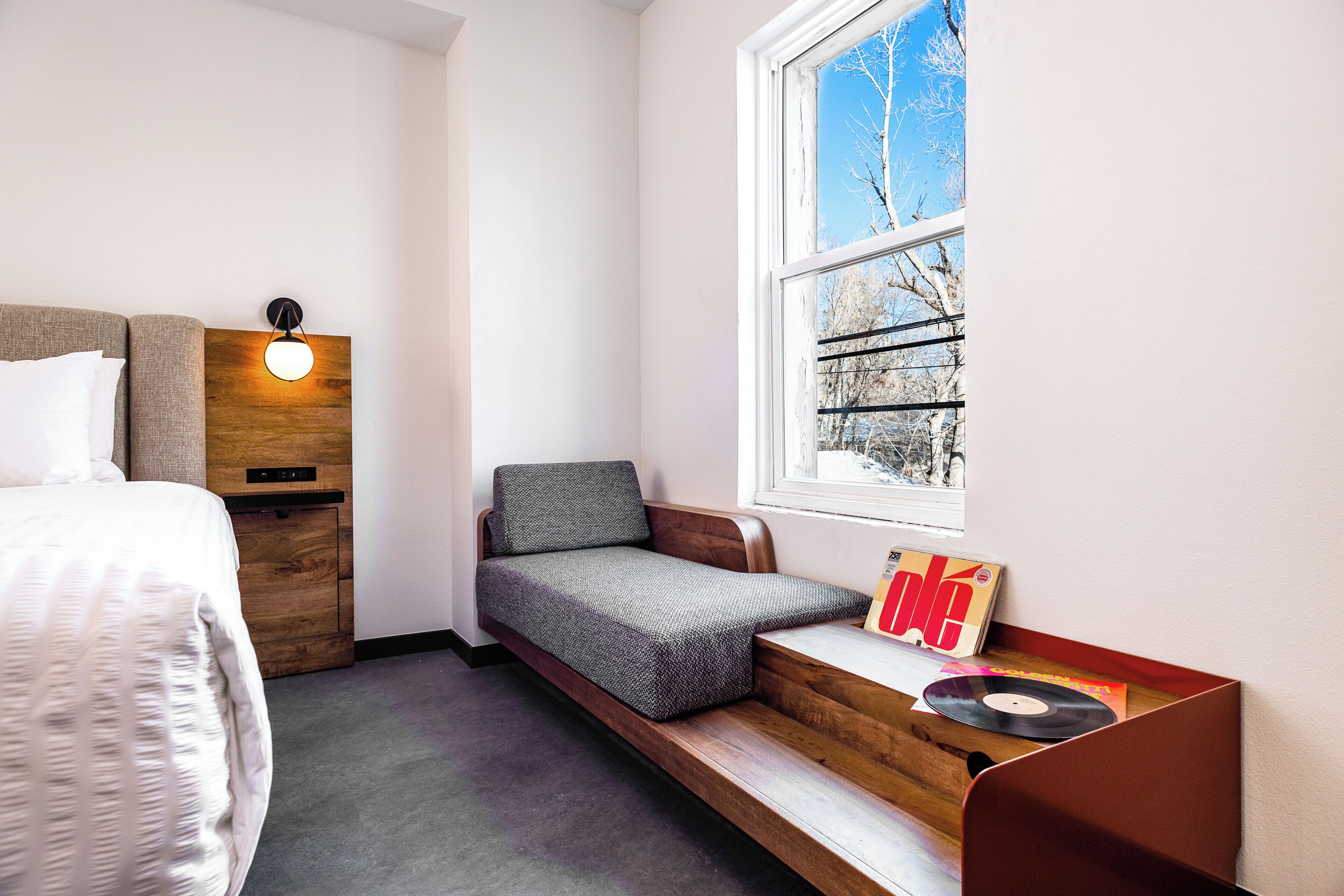 A bedroom with a bed, a built-in bench with a cushion, a bedside table with a record player, and a window with a view of trees.