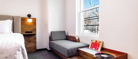 A bedroom with a bed, a built-in bench with a cushion, a bedside table with a record player, and a window with a view of trees.