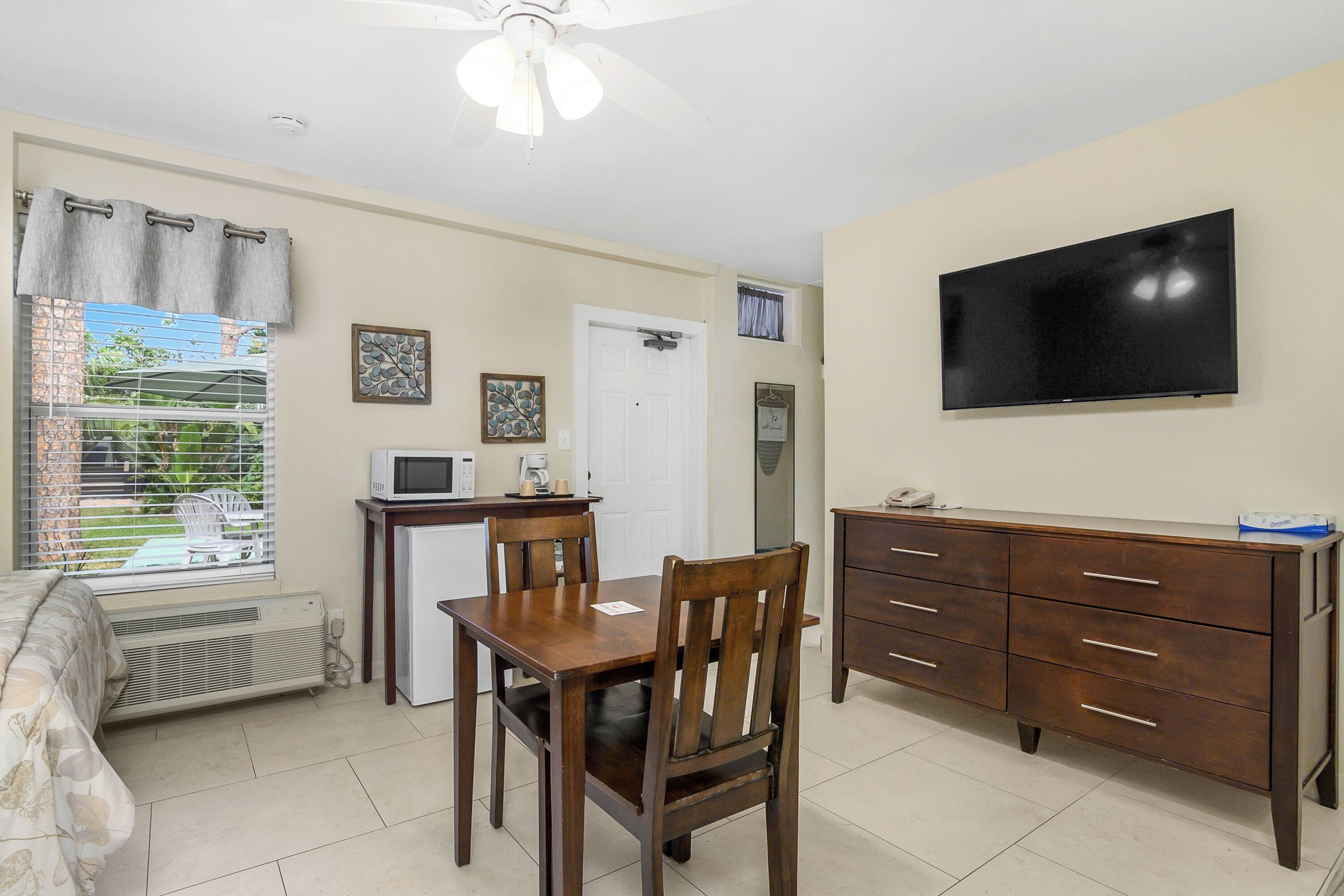 Una habitación compacta con una mesa de comedor, sillas, un televisor montado en la pared, una kitchenette pequeña y una ventana con persianas.