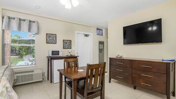 Una habitación compacta con una mesa de comedor, sillas, un televisor montado en la pared, una kitchenette pequeña y una ventana con persianas.