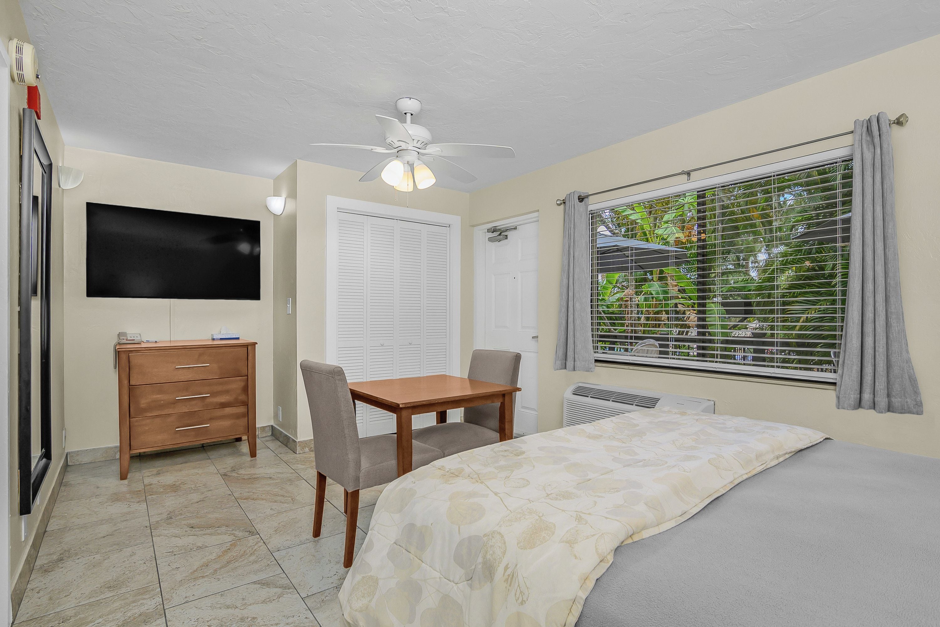 A bedroom with a bed, a desk, a chair, a television, and a window with blinds.