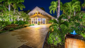 A well-lit building with a sloped roof, surrounded by lush greenery and palm trees.