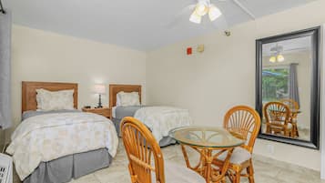 A hotel room with two beds, a small table with chairs, a ceiling fan, a large mirror, and a window with blinds.