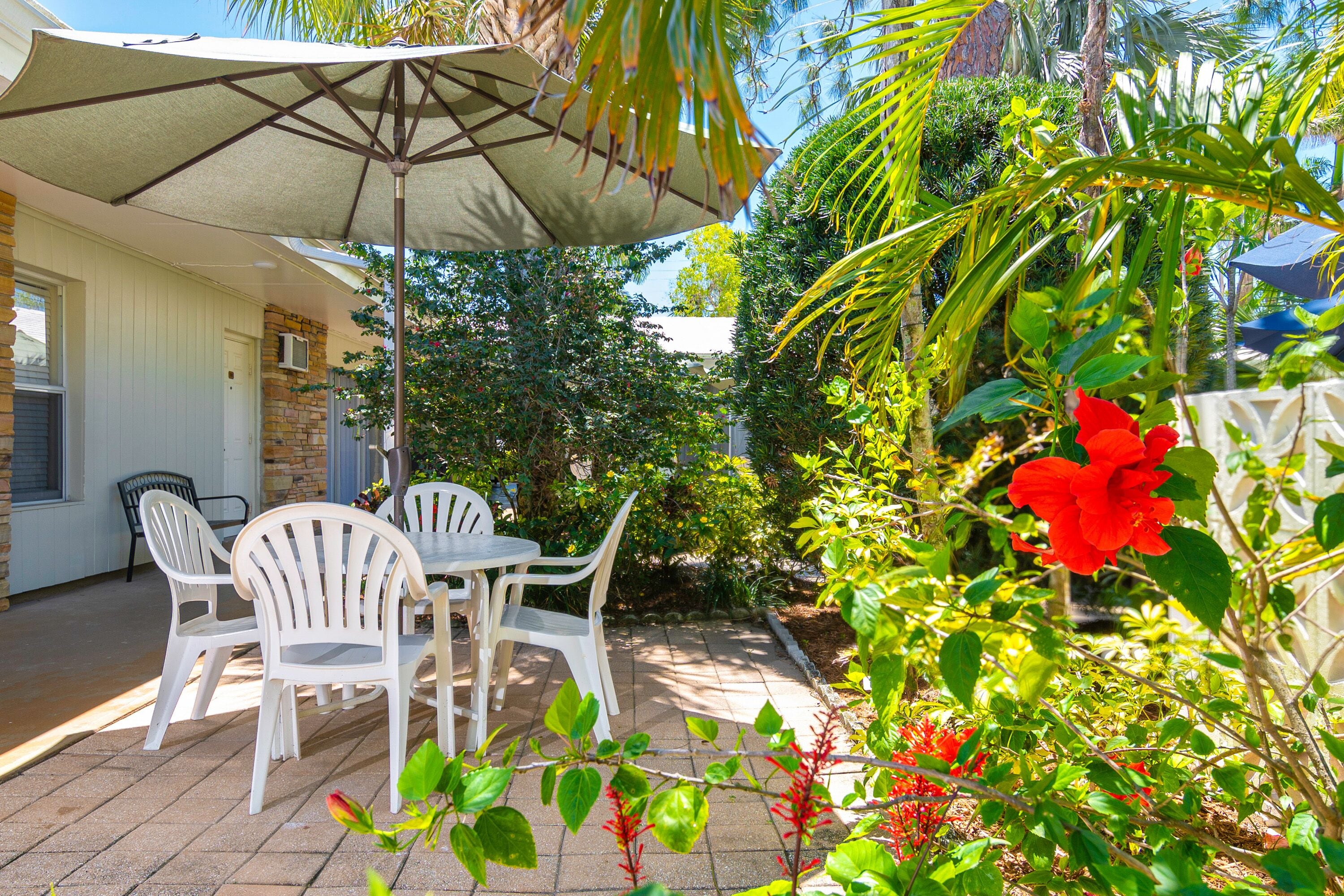 A patio with a table and chairs, surrounded by lush greenery and a red flower.