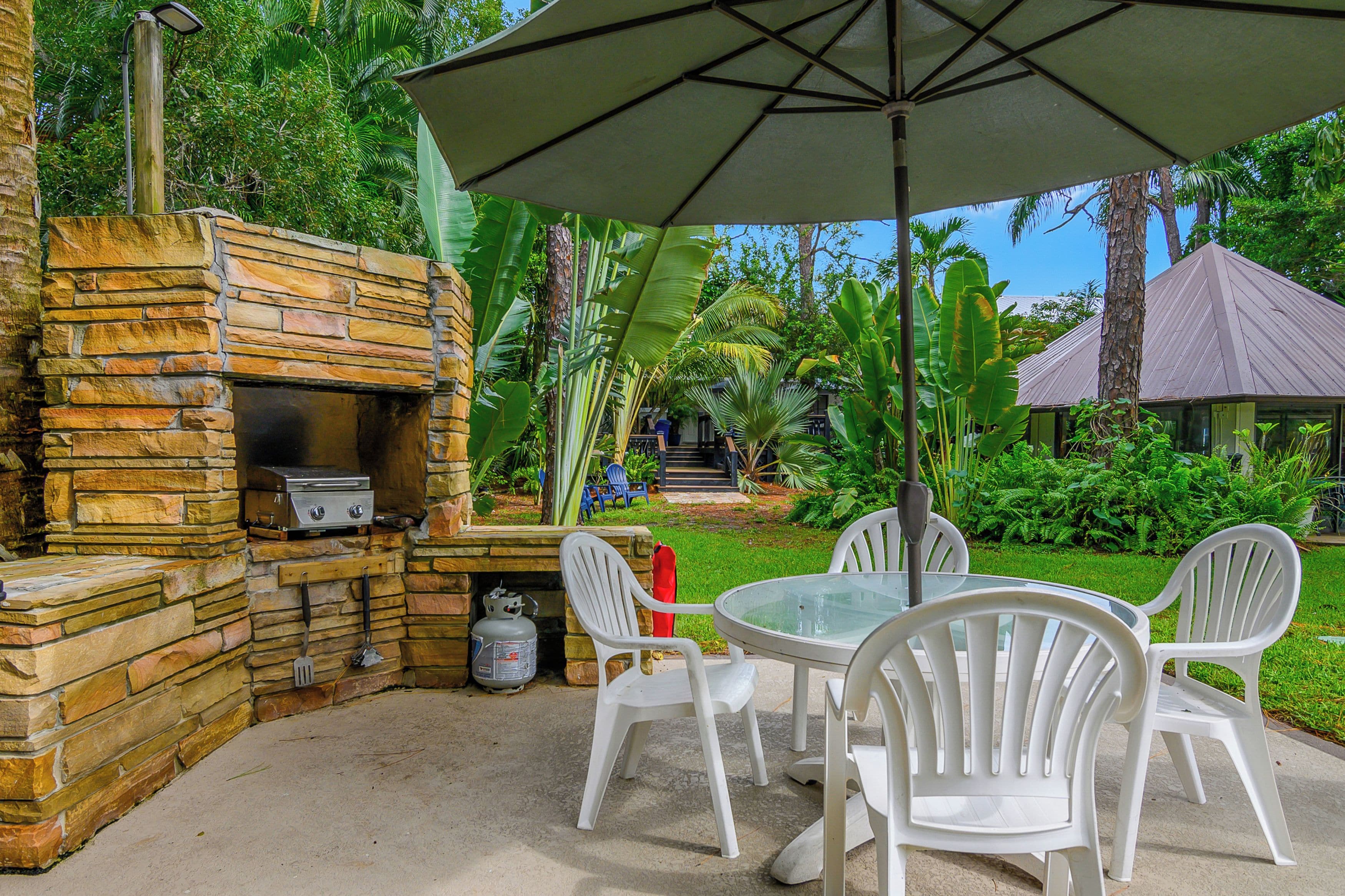 An outdoor patio with a stone barbecue grill, white plastic chairs, and a table under a large umbrella.
