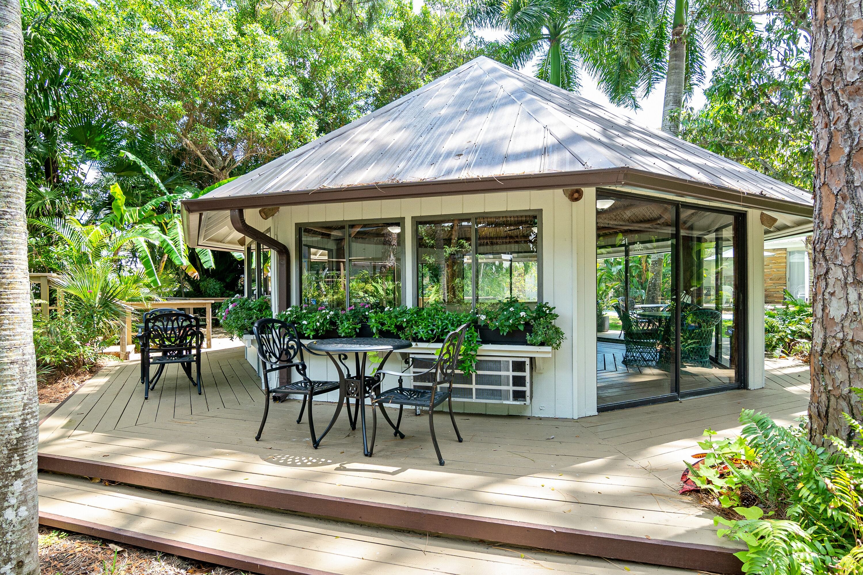 A covered outdoor seating area with a table and chairs, surrounded by lush greenery.