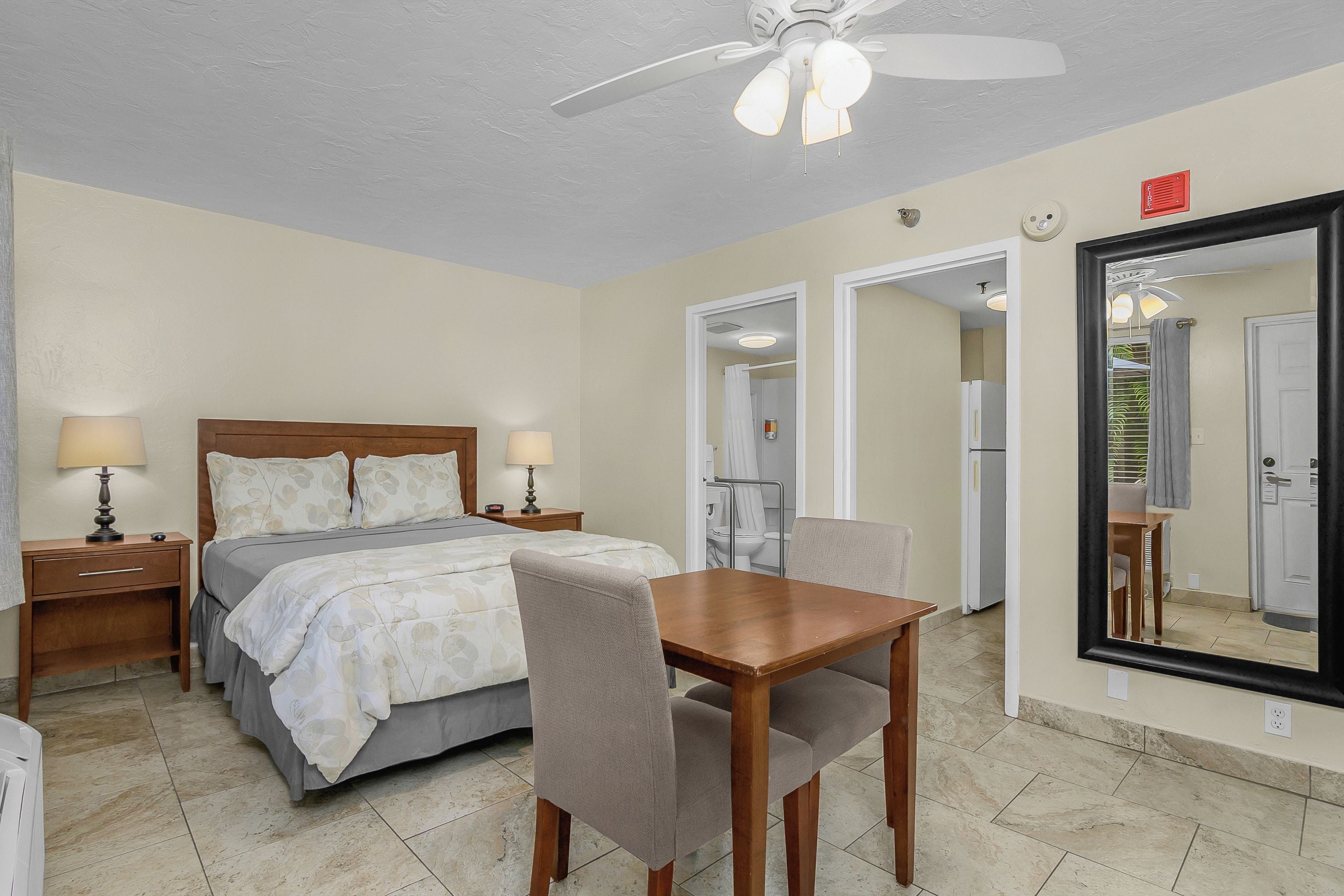 A hotel room with a bed, bedside tables, a desk with a chair, a ceiling fan, and a large mirror.