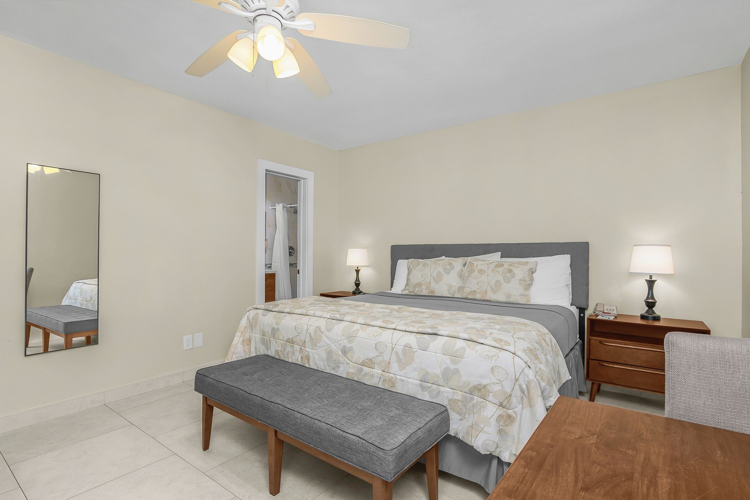 Une chambre à coucher comprenant un lit, des tables de chevet, un banc, un ventilateur de plafond et un miroir.