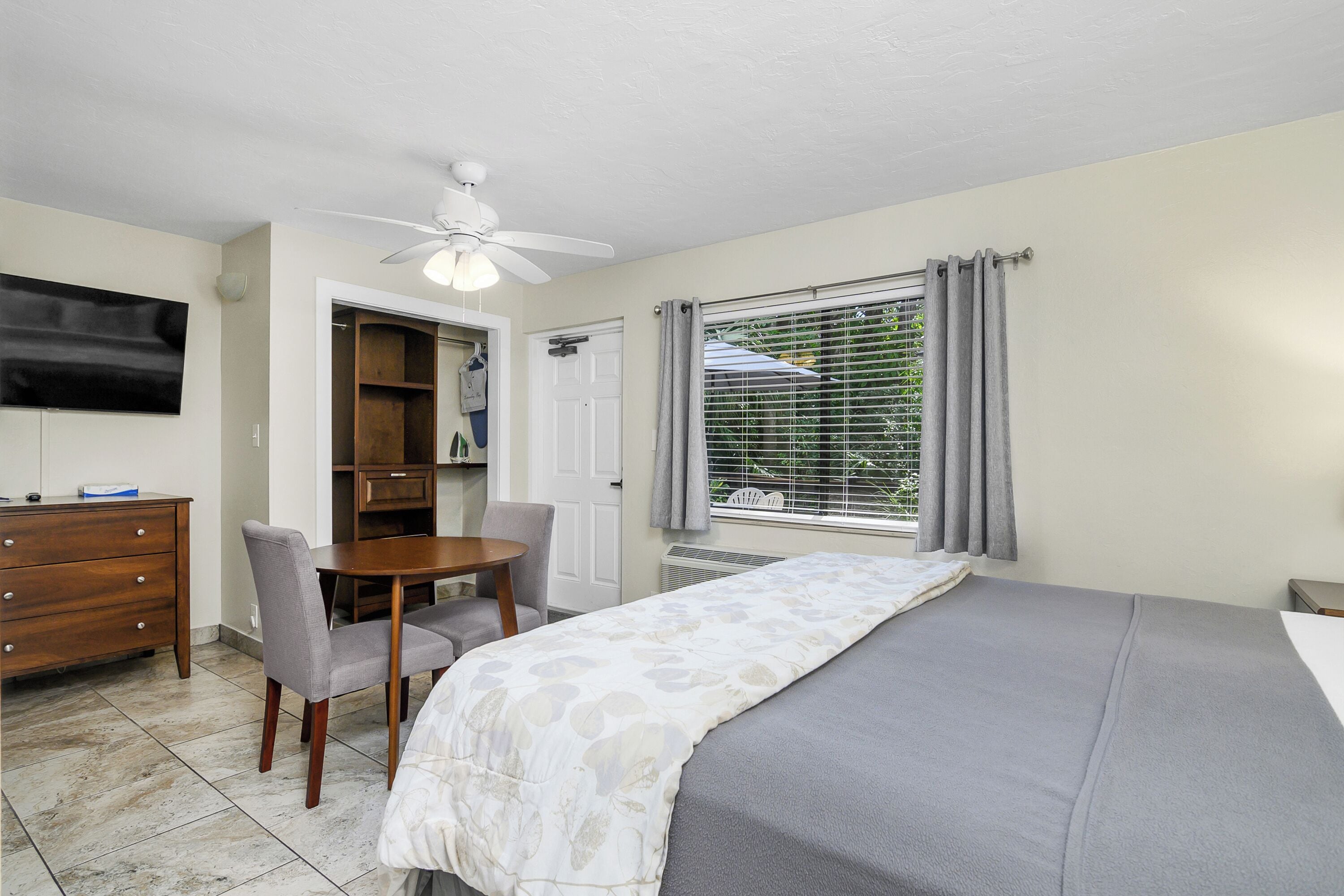 A bedroom with a bed, a desk, a chair, a ceiling fan, a TV, and a window with curtains.