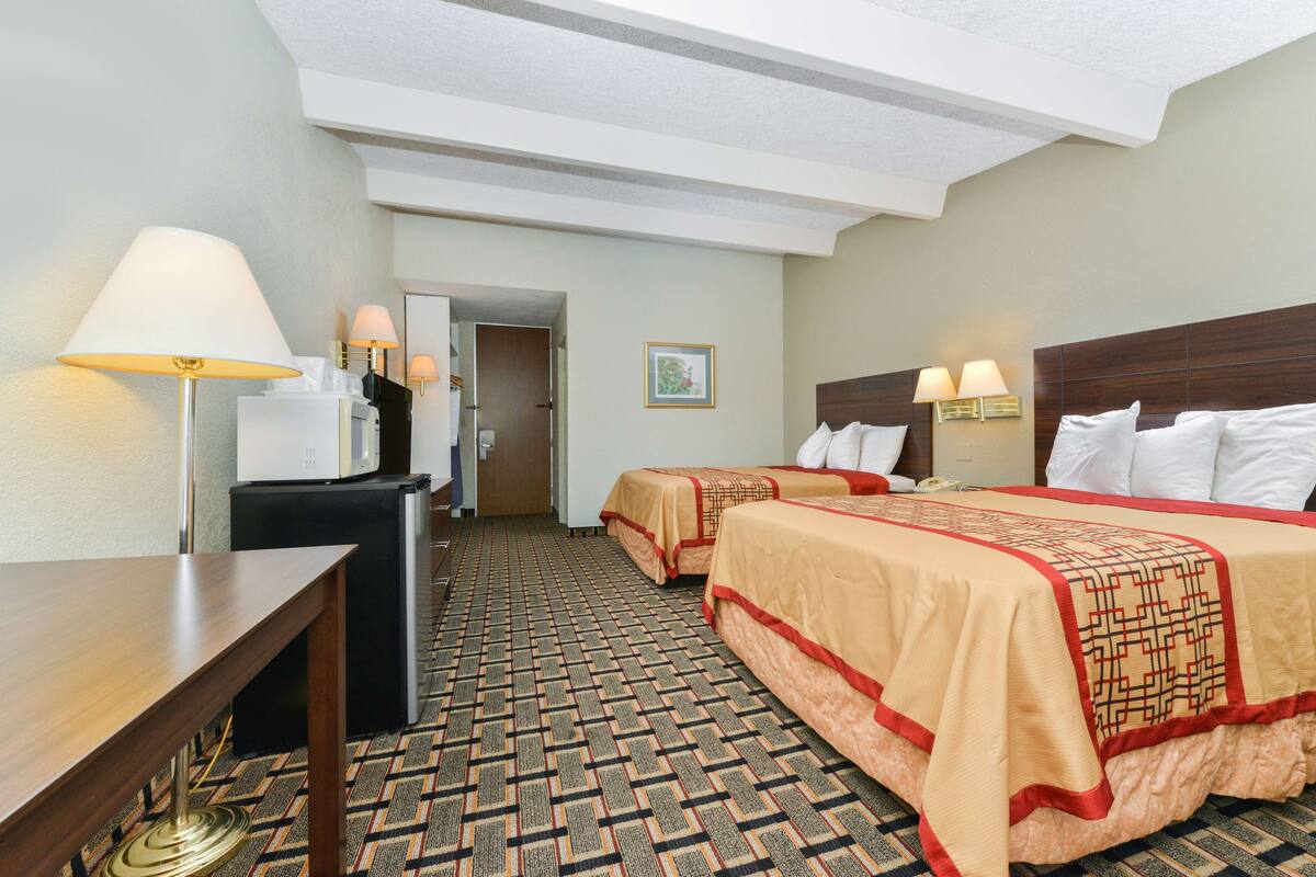 A hotel room with two beds, a desk, a refrigerator, and a lamp.