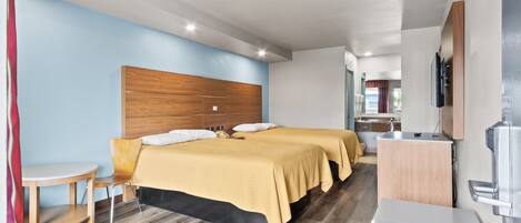 Habitación de hotel moderna con cabecera de madera, dos camas con ropa de cama amarilla, una mesita redonda y un televisor montado en la pared.