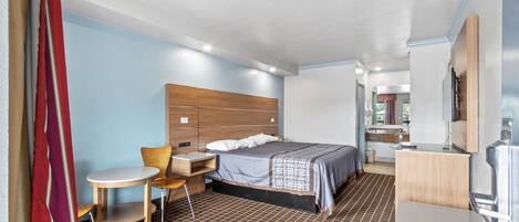 A hotel room with a bed, a desk, a chair, a small table, and a kitchenette.