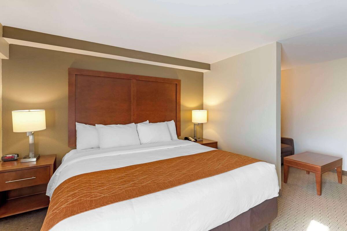 Habitación de hotel con una cama grande, dos mesitas de noche con lámparas, un cabecero de madera y una pequeña mesita auxiliar de madera.