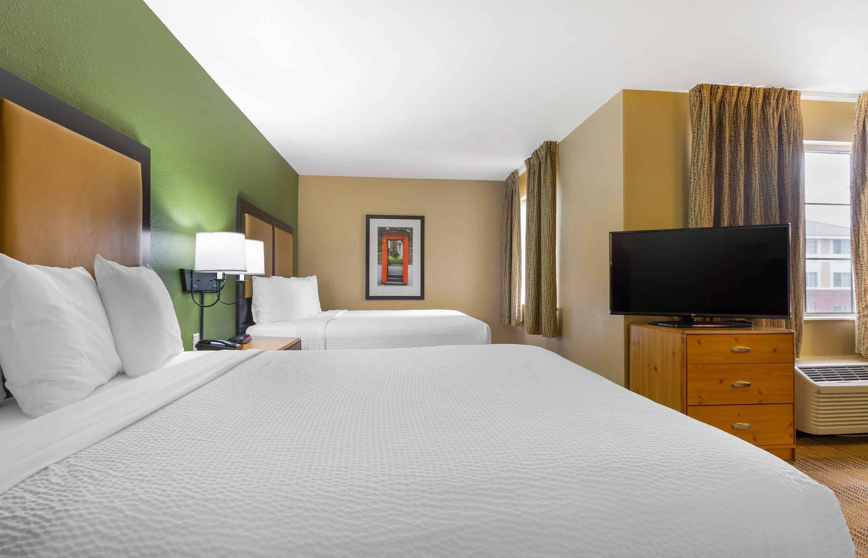 Quarto de hotel com duas camas, televisão, mesa de cabeceira com abajur, quadro na parede e janela com cortinas.