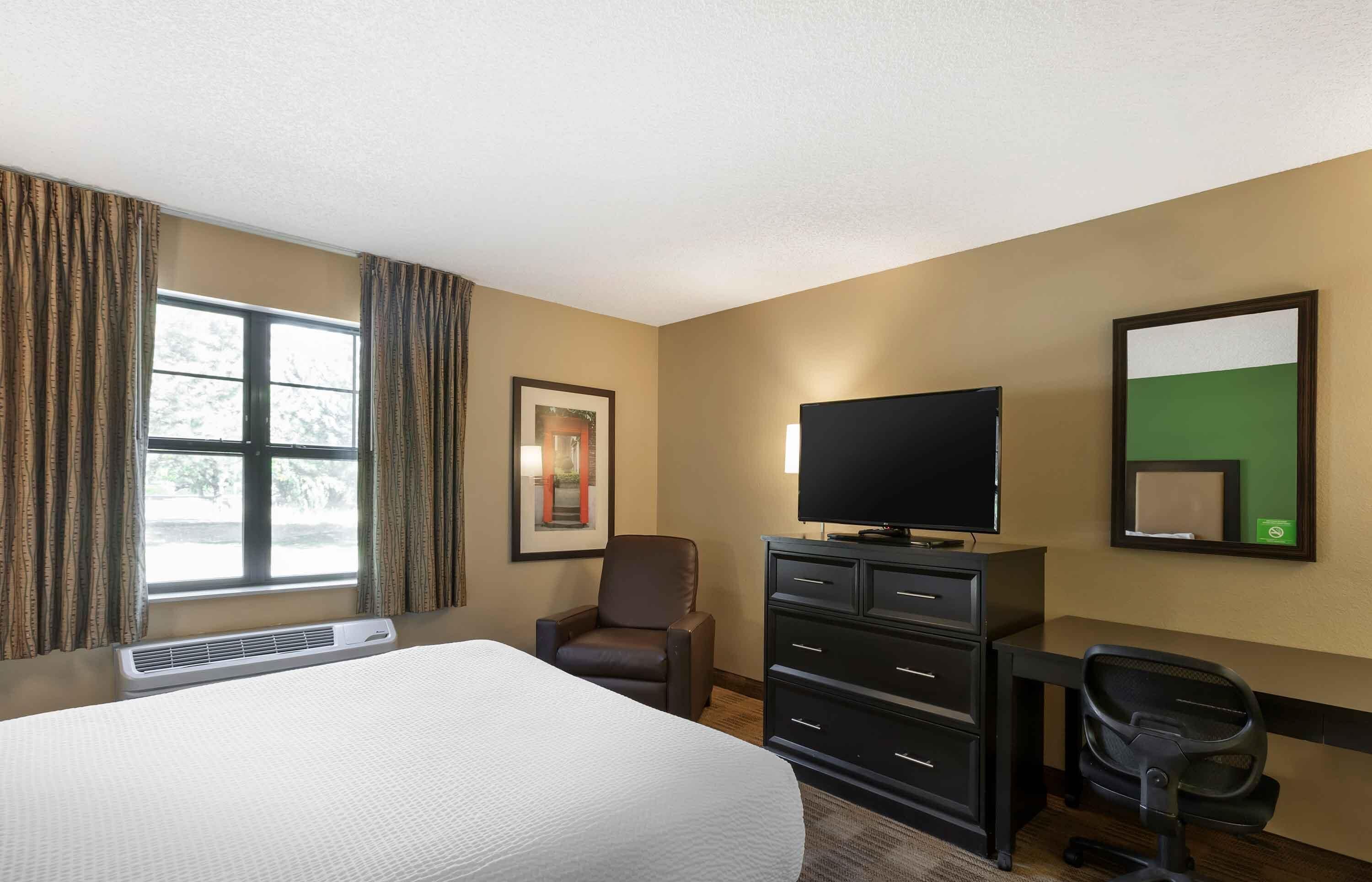 Quarto de hotel com uma cama, uma secretária com televisão de tela plana, uma cadeira, uma luminária de mesa e um quadro na parede.