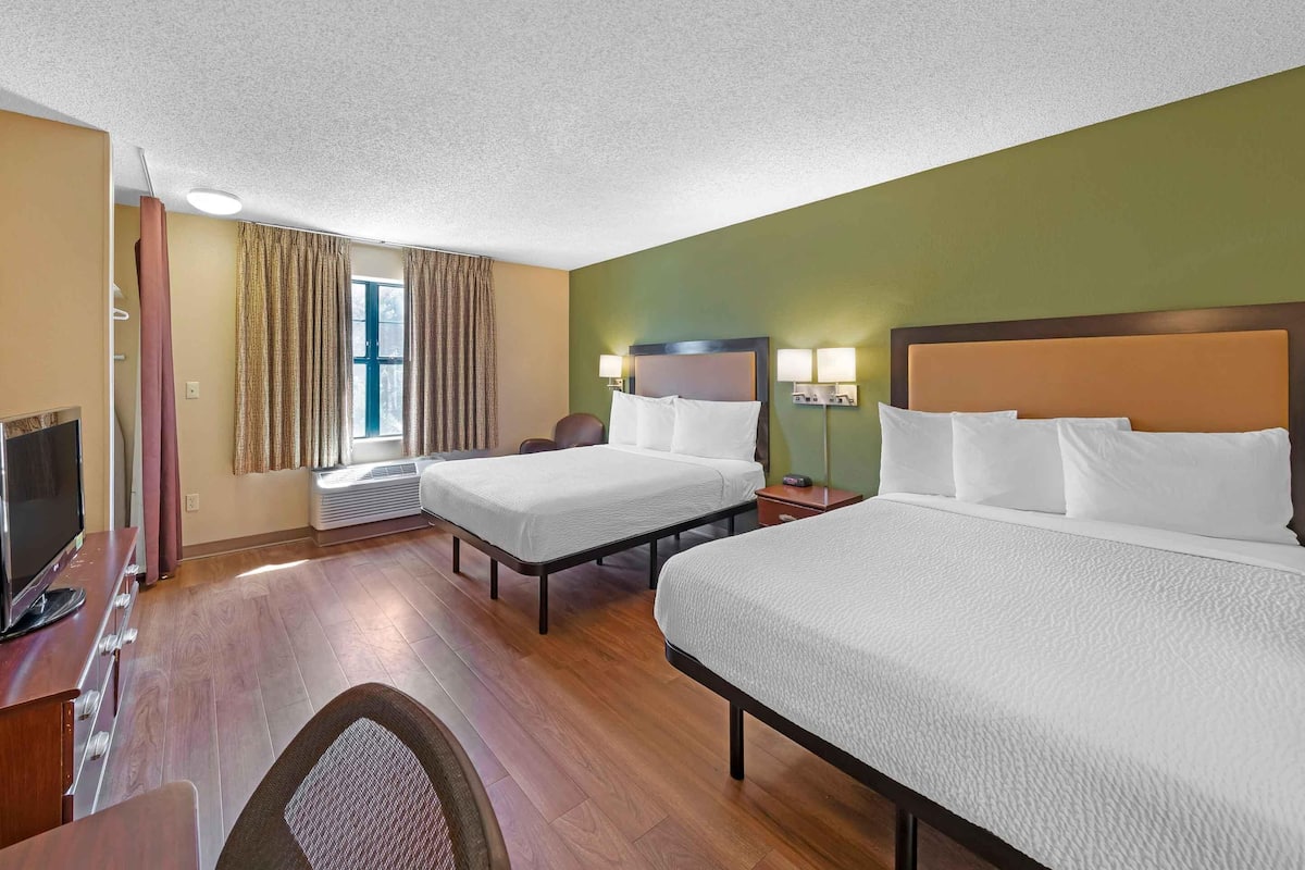 A hotel room with two beds, a desk with a television, a chair, and a window with curtains.