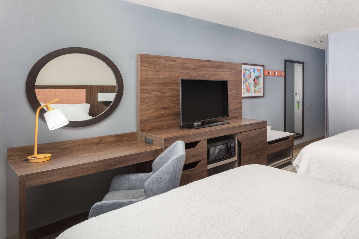 A hotel room with a wooden desk, a flat-screen TV, a mirror, a bed, and a door.