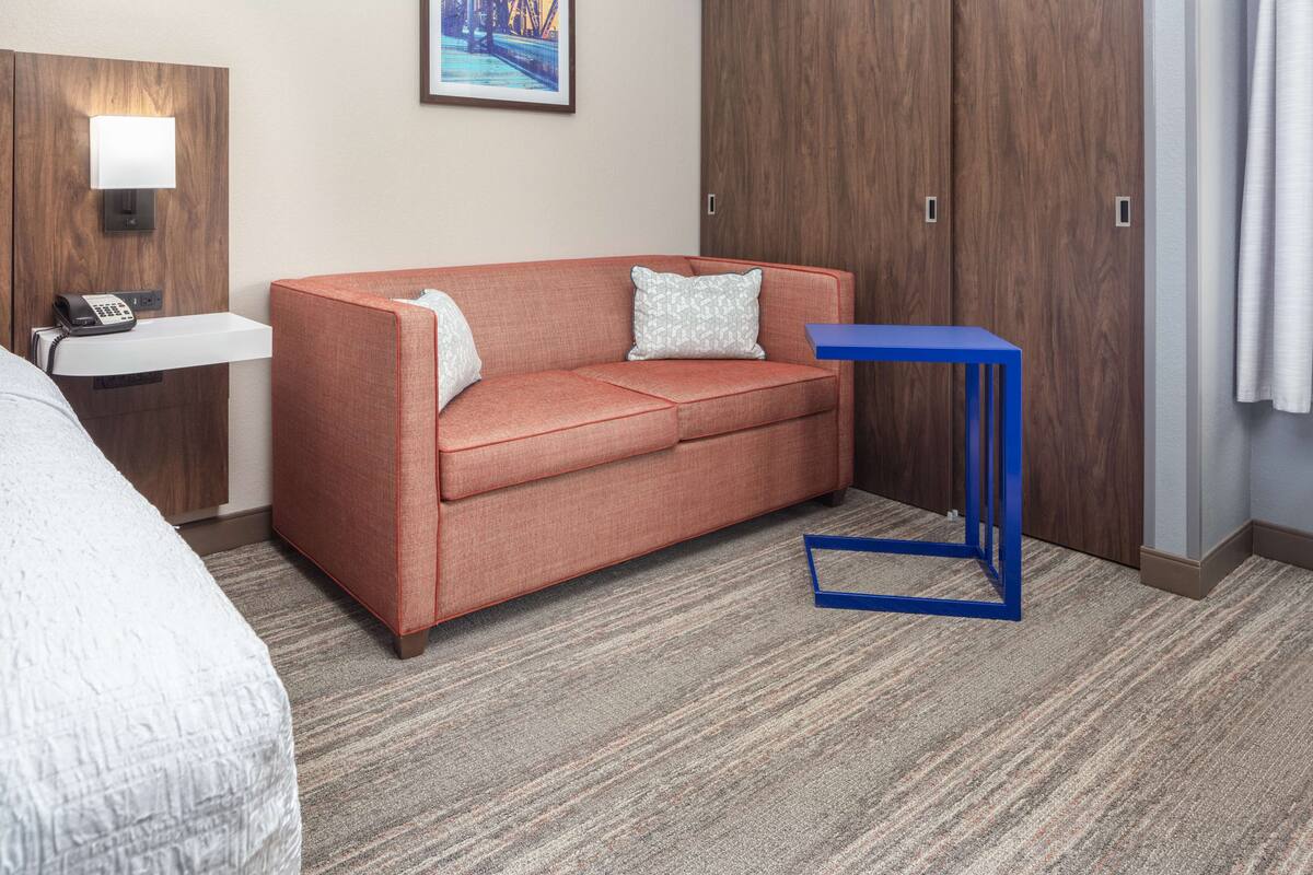 A hotel room with a red sofa, a small table, a bedside table with a lamp, and a wardrobe.