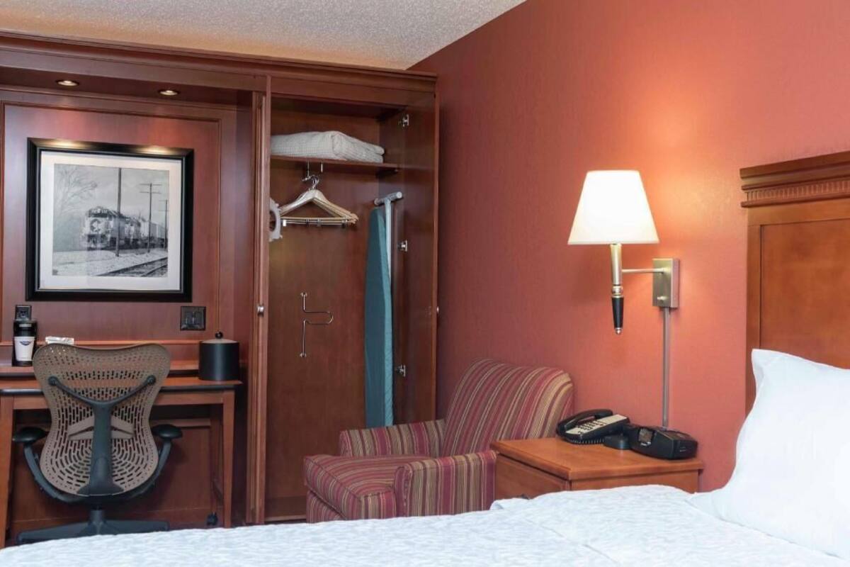 A hotel room with a bed, a desk with a chair, a hanging coat rack, a shower, and a bedside lamp.