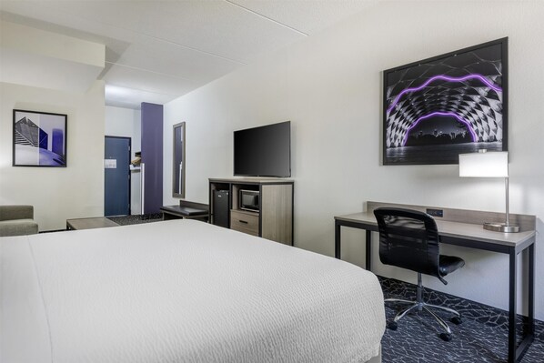 A hotel room with a large bed, a desk with a chair, a flat-screen TV, a desk lamp, and a framed artwork on the wall.