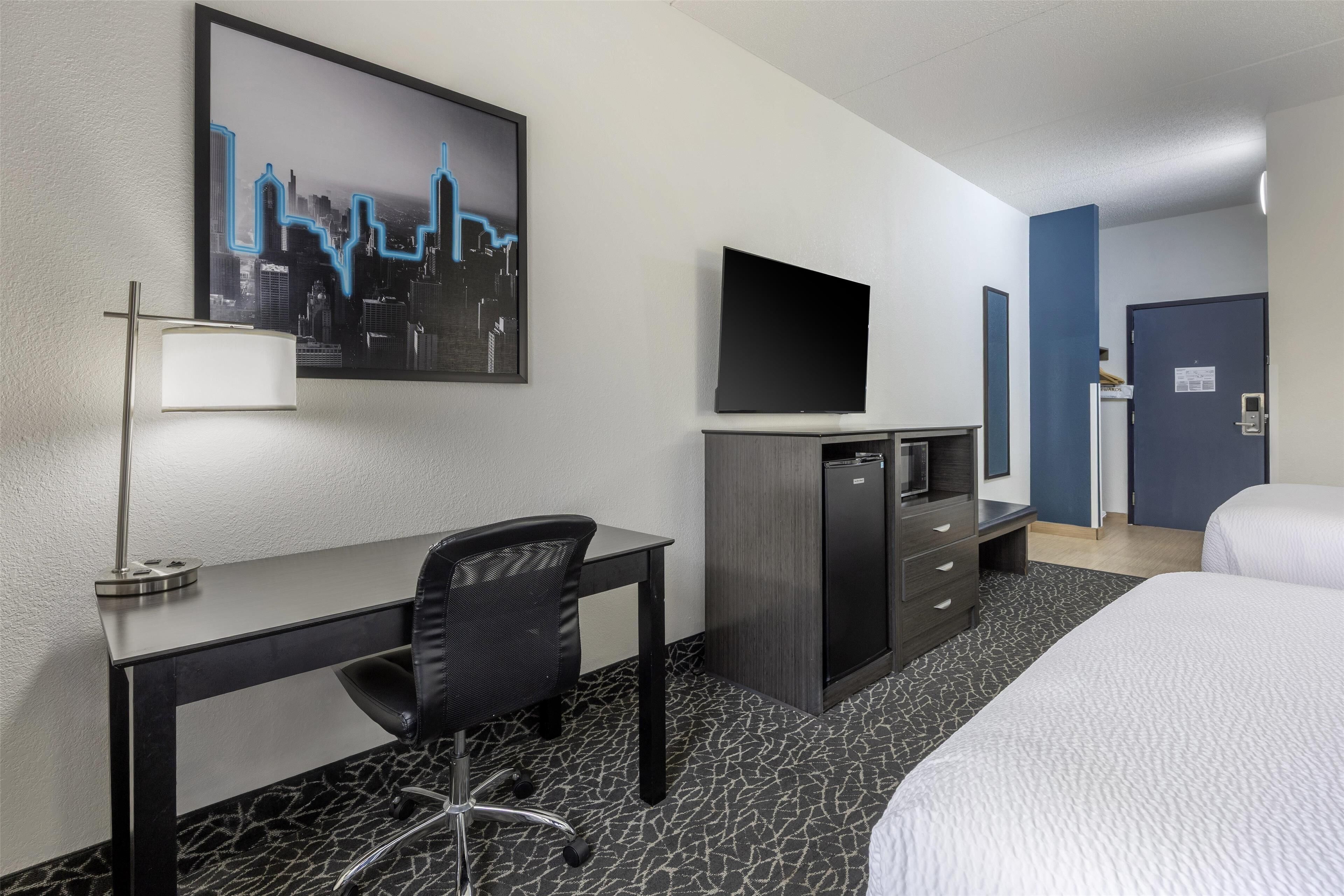 A hotel room with a bed, a desk with a chair, a TV, a dresser, and a cityscape painting on the wall.