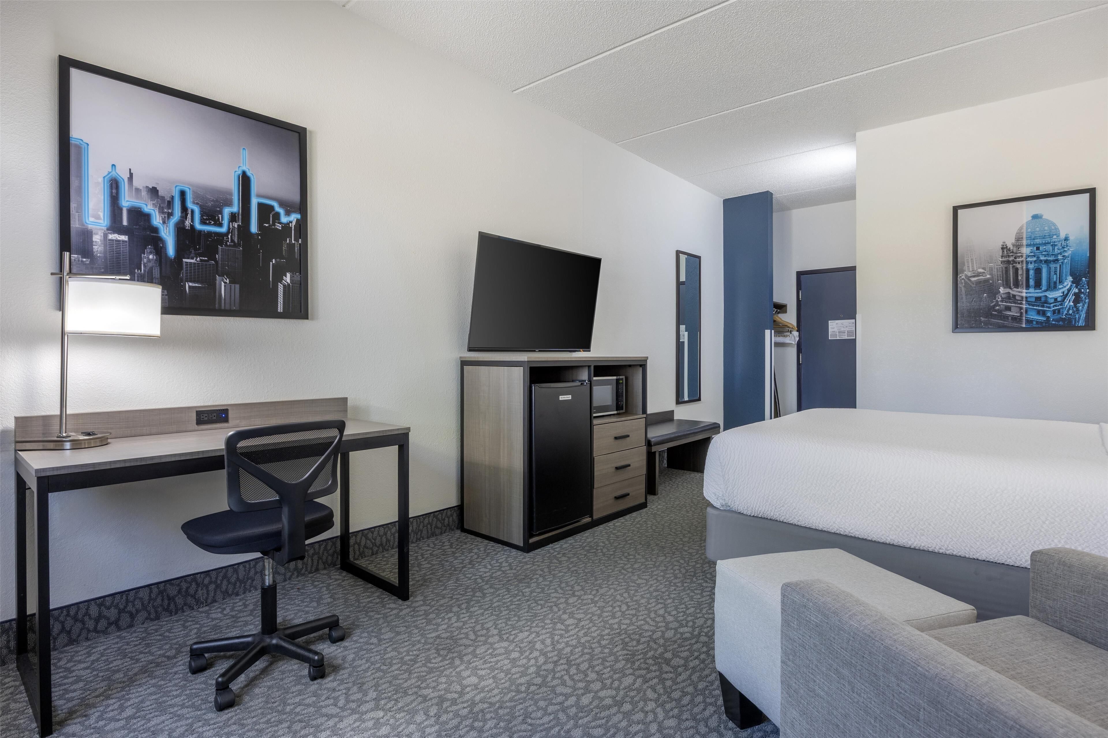 A hotel room with a bed, a desk with a computer, a chair, a TV, and artwork on the walls.