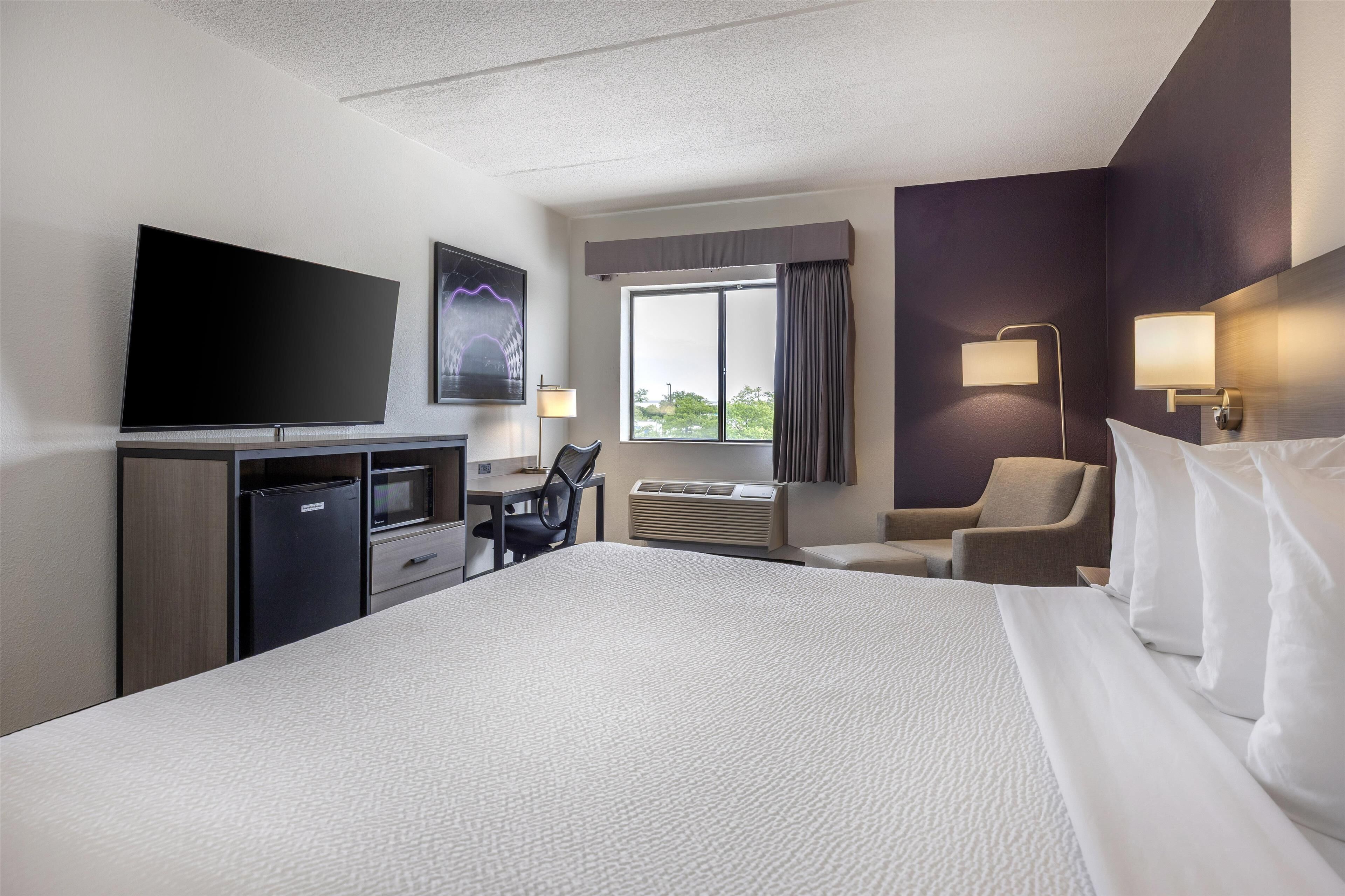 Habitación de hotel con una cama grande, un escritorio con televisor de pantalla plana, una silla, una lámpara de escritorio y un ventanal con cortinas.