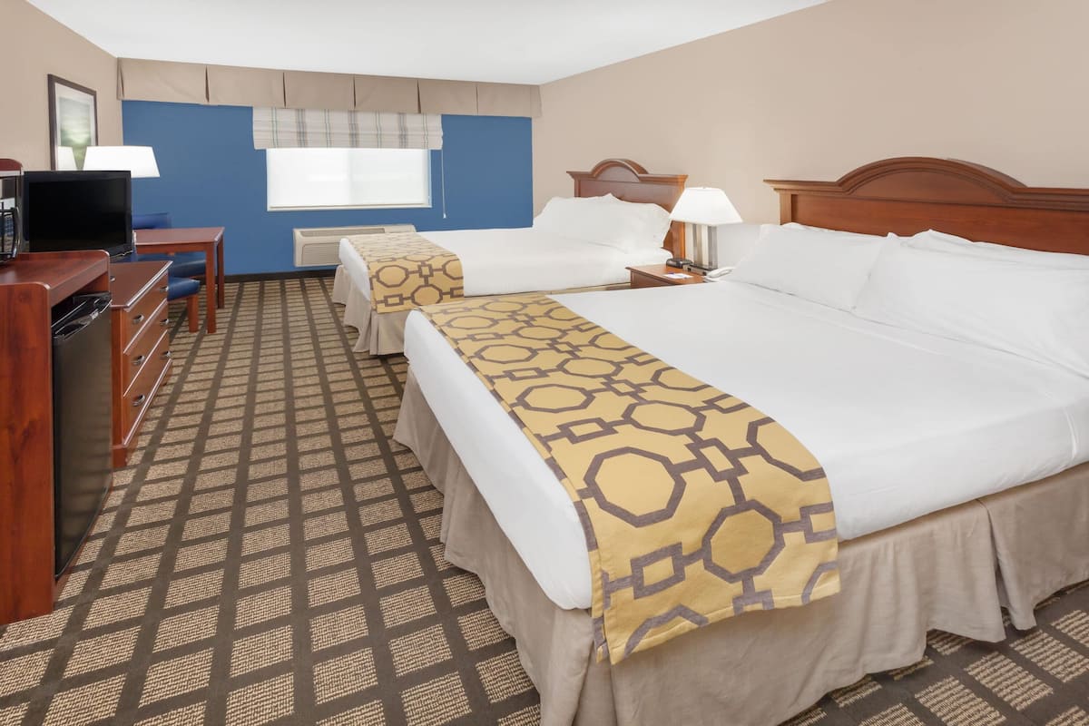 Une chambre d’hôtel avec un grand lit, un plus petit lit, un bureau avec une chaise, une télévision et une fenêtre avec des stores.