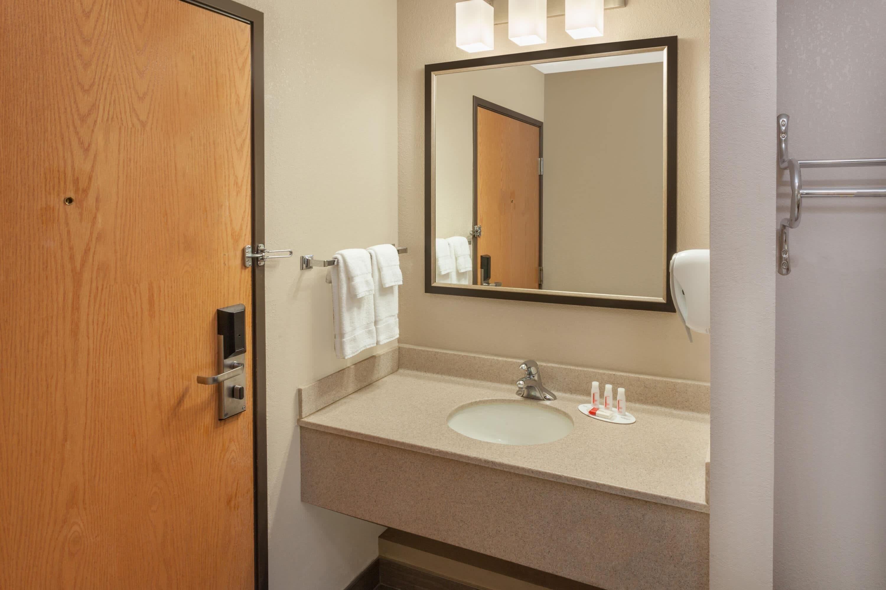 Un baño con puerta de madera, un lavabo con espejo y un perchero con toallas blancas.