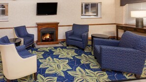 A hotel room with a fireplace, two blue armchairs, a sofa, a flat-screen TV, and a window with blinds.