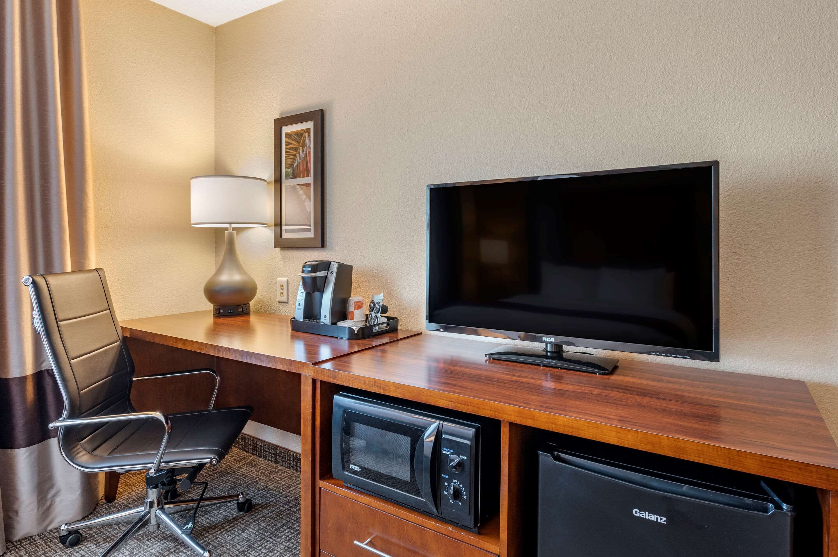 Quarto de hotel com escrivaninha de madeira, televisão, micro-ondas, cafeteira e uma cadeira.