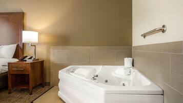 Un baño de hotel con una bañera blanca grande, una mesita de noche con una lámpara y un perchero en la pared.