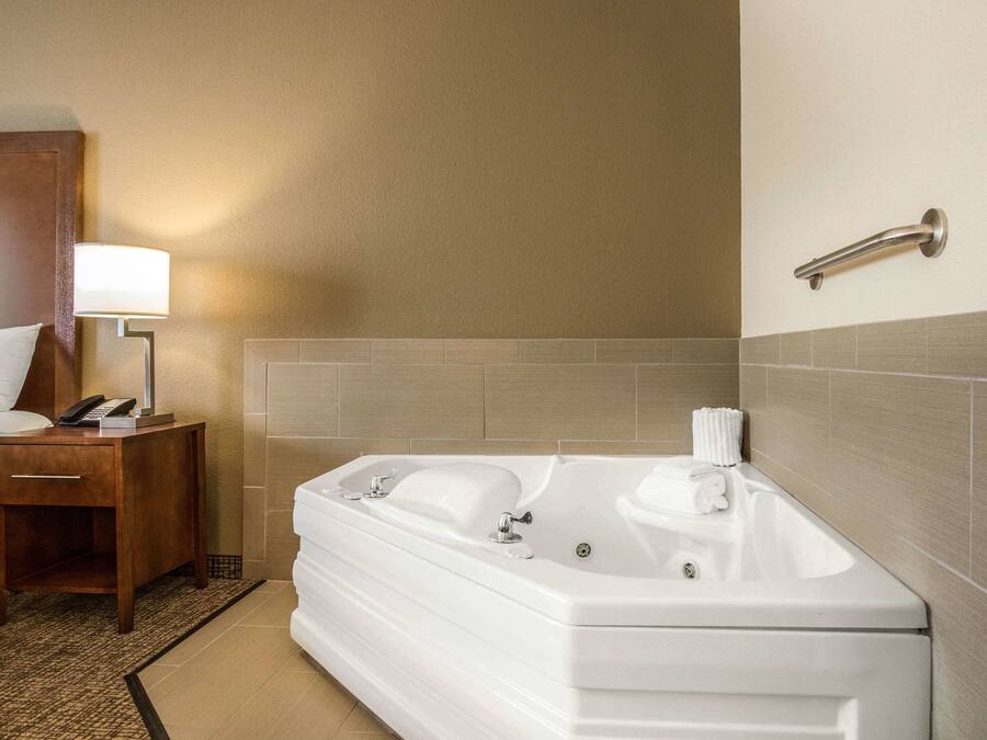 Un baño de hotel con una bañera blanca grande, una mesita de noche con una lámpara y un perchero en la pared.