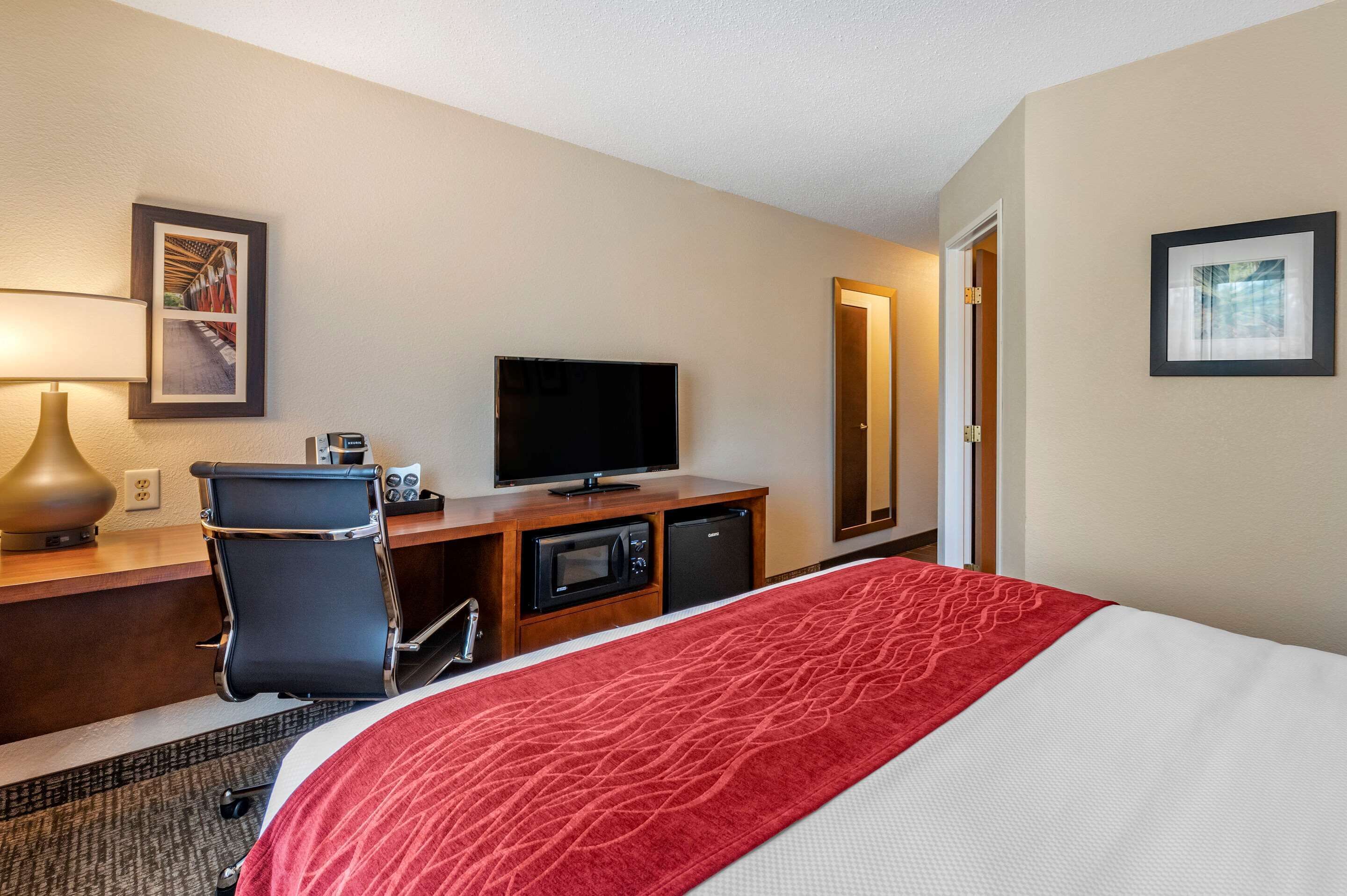 A hotel room with a bed, a desk with a television, a chair, a lamp, and a microwave.