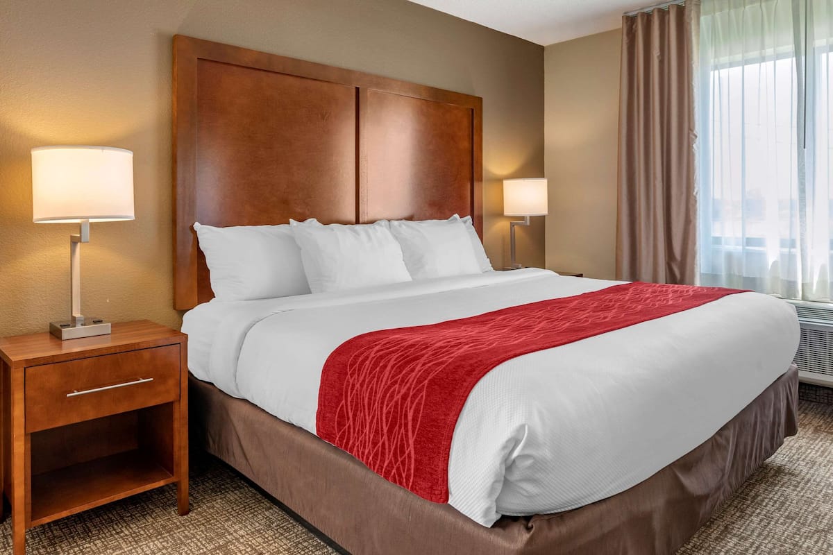 Ein Hotelzimmer mit einem großen Bett, zwei Nachttischlampen, einem Holz-Kopfstück und einem Fenster mit Vorhängen.