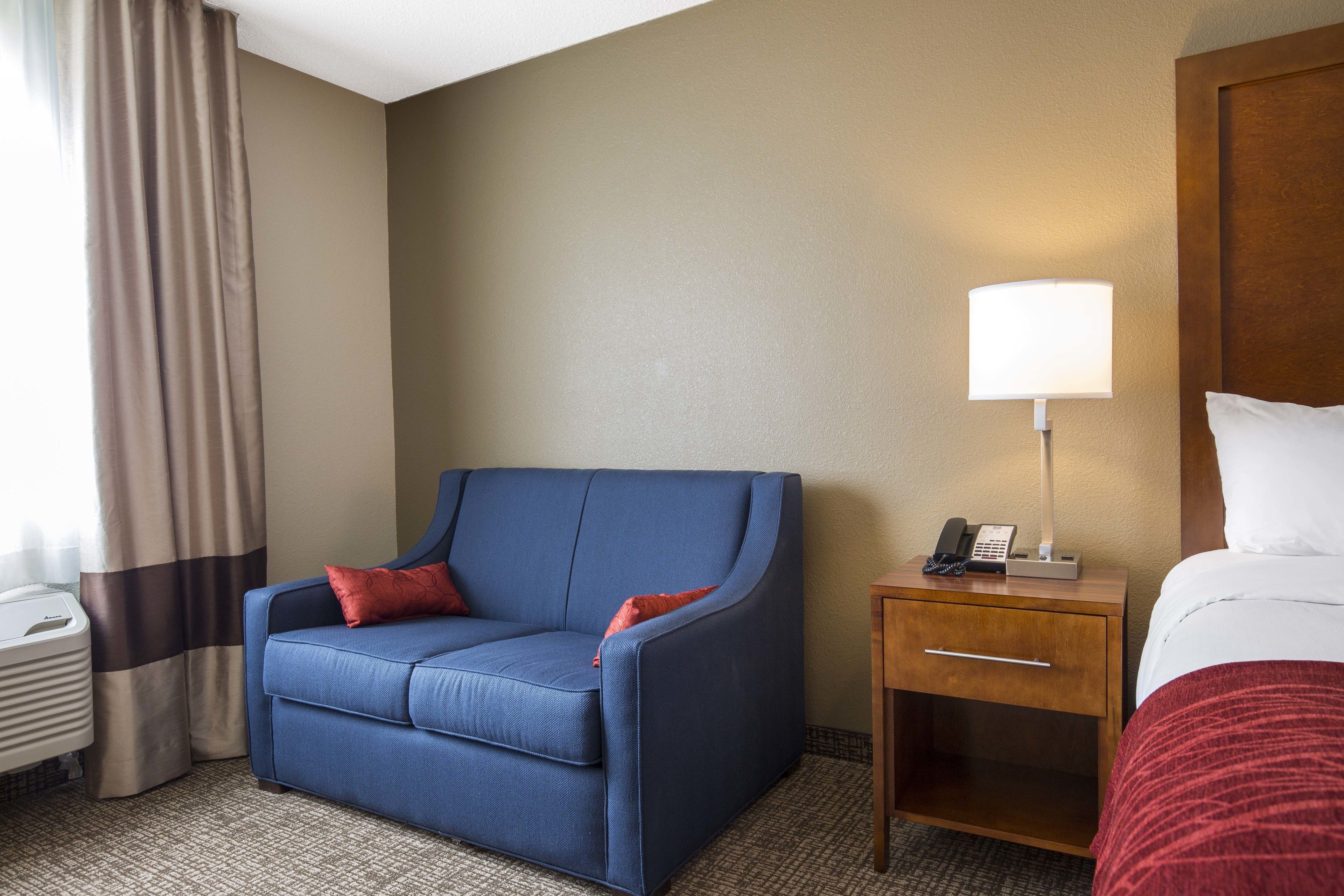 A hotel room with a blue sofa, a wooden nightstand with a lamp, a telephone, and a bed with white bedding.