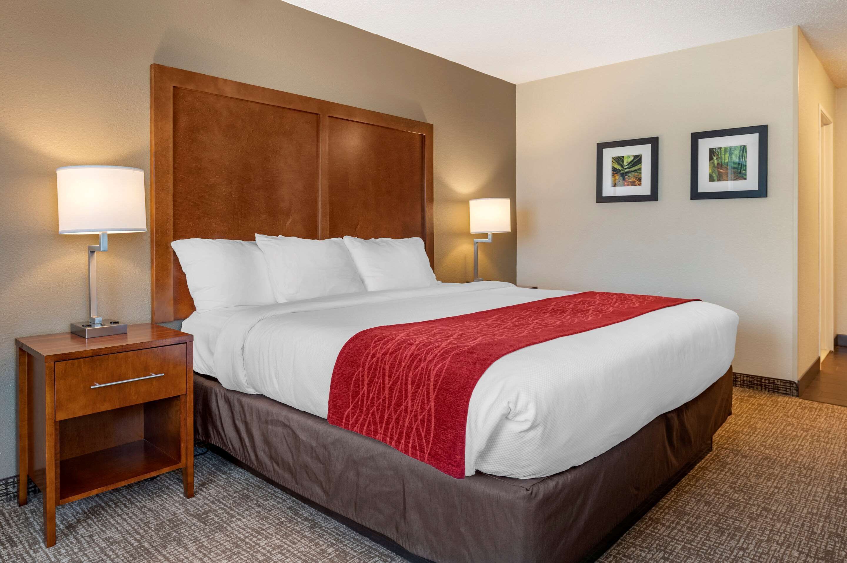 A hotel room with a large bed, wooden headboard, two bedside lamps, a nightstand, and two framed pictures on the wall.