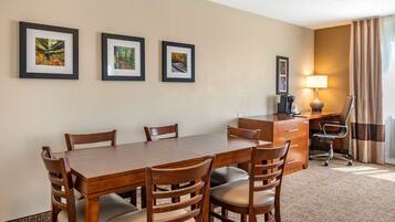 A hotel room with a wooden dining table, six chairs, a desk with a chair, a lamp, and three framed pictures on the wall.