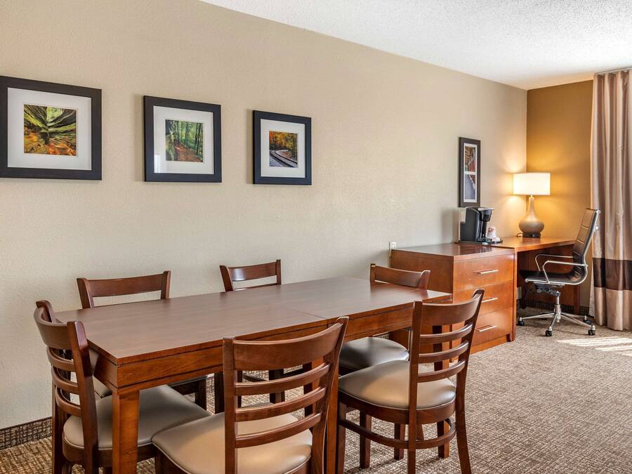 A hotel room with a wooden dining table, six chairs, a desk with a chair, a lamp, and three framed pictures on the wall.