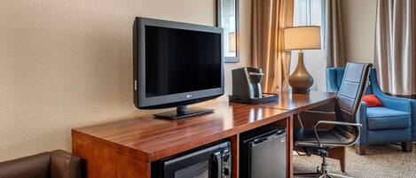 A hotel room with a wooden desk, a flat-screen TV, a microwave, a mini-fridge, a blue armchair, and a lamp.