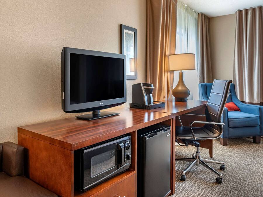 A hotel room with a wooden desk, a flat-screen TV, a microwave, a mini-fridge, a blue armchair, and a lamp.
