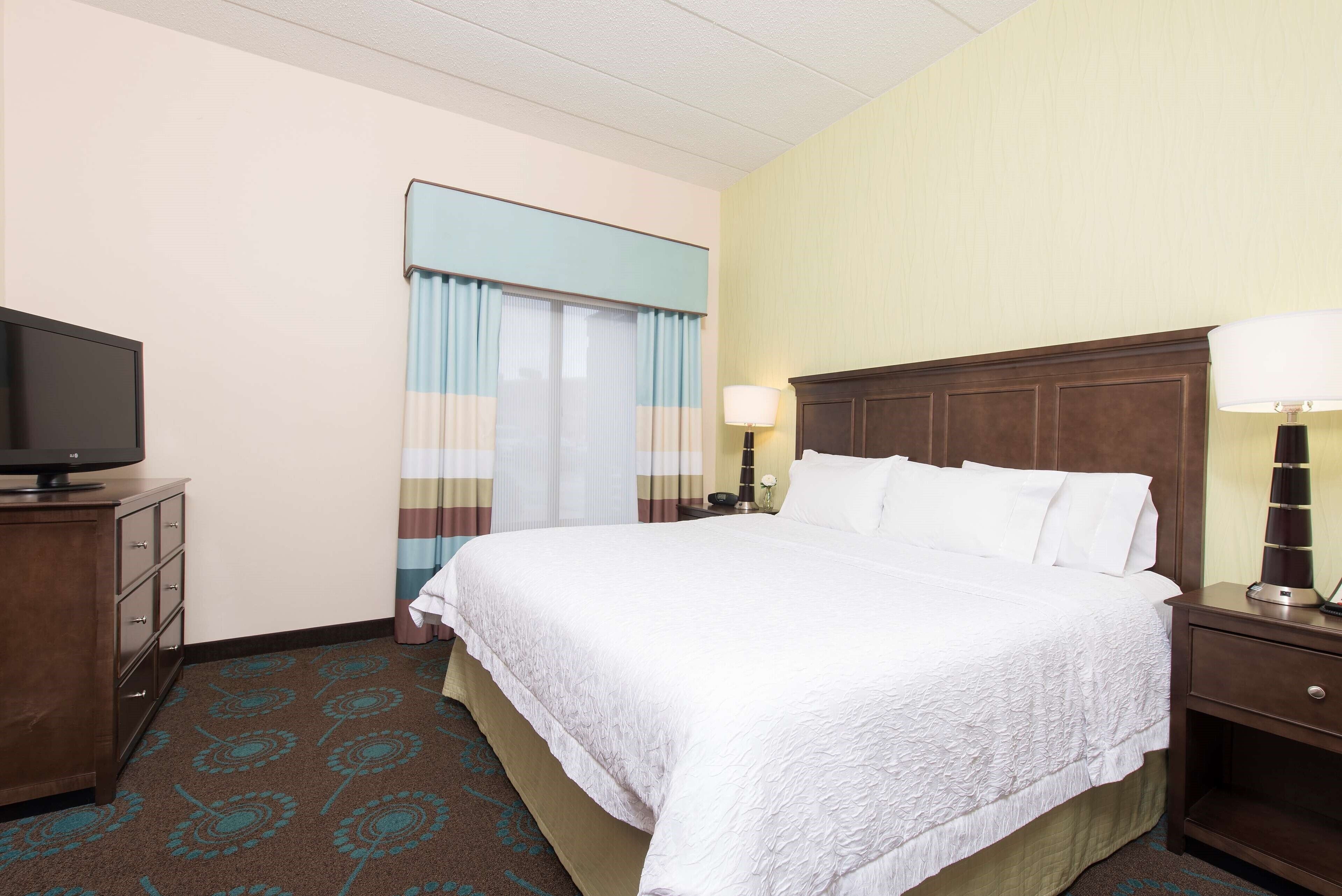 Una habitación de hotel con una cama grande, un televisor sobre una cómoda de madera y una mesita de noche con una lámpara.