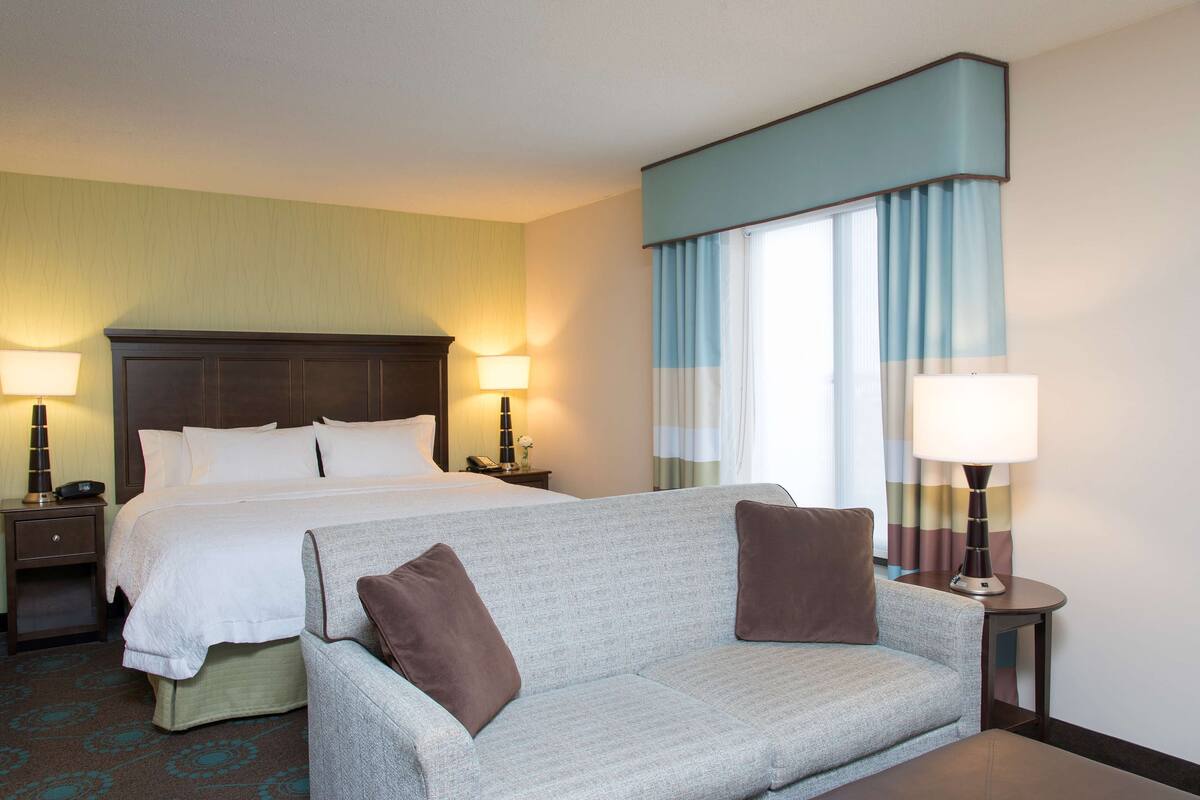 A hotel room with a bed, a sofa, two bedside tables with lamps, and a window with curtains.