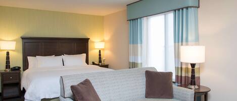 A hotel room with a bed, a sofa, two bedside tables with lamps, and a window with curtains.
