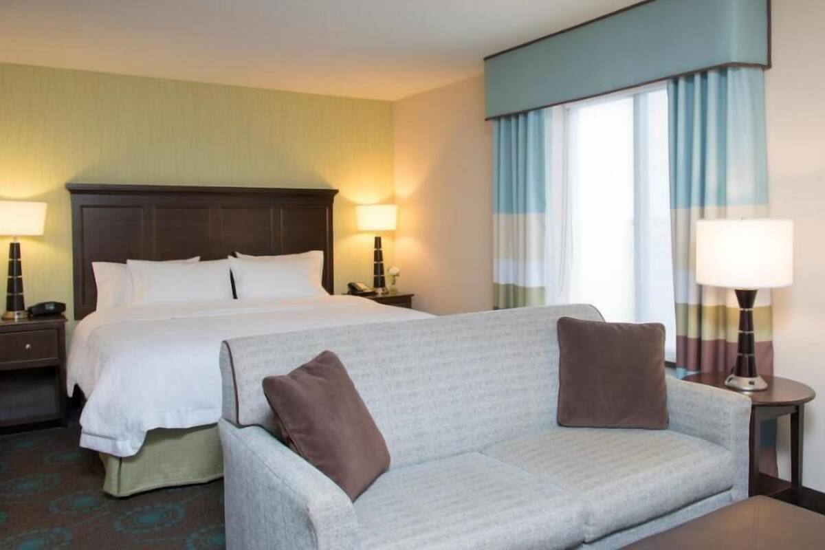 A hotel room with a double bed, a grey sofa, two bedside lamps, a nightstand, and a window with curtains.