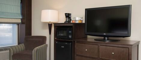 A hotel room with a bed, a brown armchair, a desk with a flat-screen TV, a microwave, a coffee maker, and a lamp.