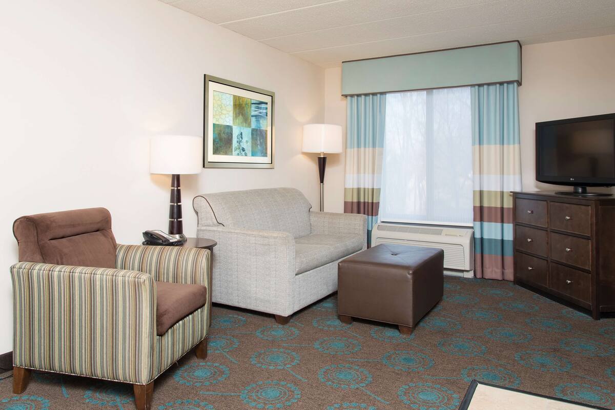 A hotel room with a striped armchair, a sofa, a small ottoman, a television, a lamp, a framed picture on the wall, and a window with curtains.